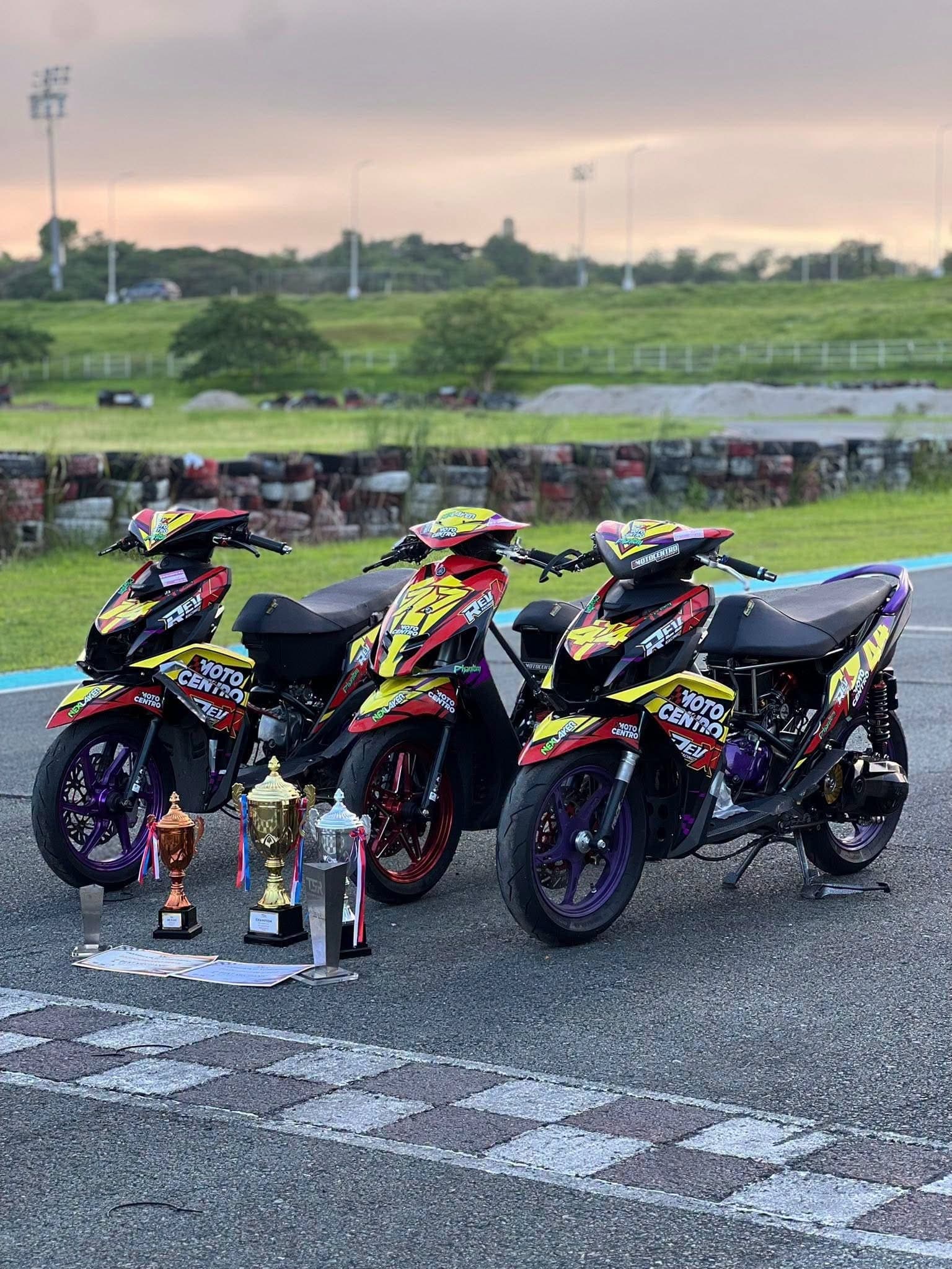 Three of the team's race bikes on the grid at dusk, with three trophies and certificates laid out in front of them